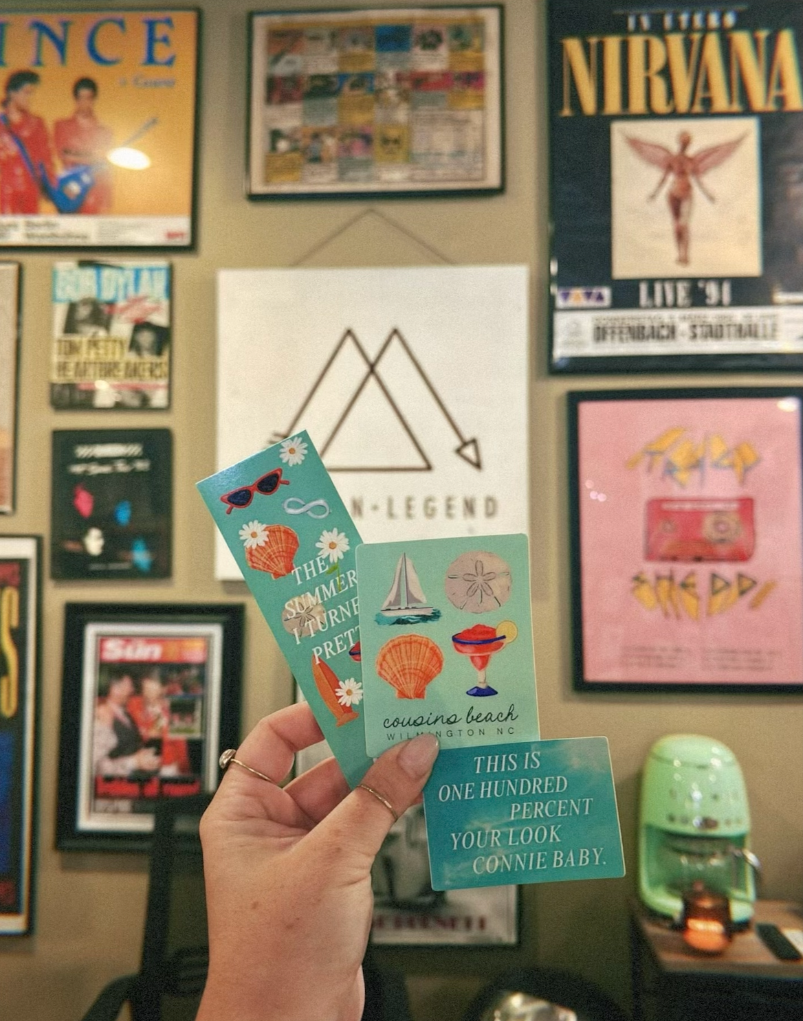 A hand is holding up one bookmark that reads, The Summer I Turned Pretty, which has a blue background with daisies, orange seashells, silver infinity signs, a surf board and red sunglasses featured. To the right of the bookmark, the hand also holds two stickers: the one closest to the bookmark depicts a sail boat, a sand dollar, an orange seashell and a strawberry daiquiri against a light teal background, and the sticker underneath that one is against a clear blue wave background and reads, in bold white letters, This is one hundred percent your look, Connie baby.