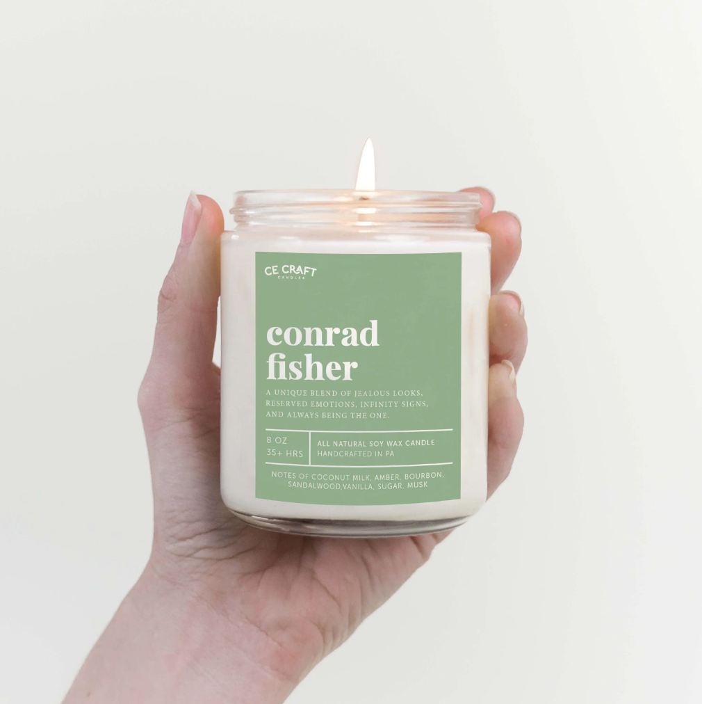 A hand is holding a lit white candle that has a green label on the front reading 'conrad fisher.' Below that, the candle's notes and ingredients are listed.